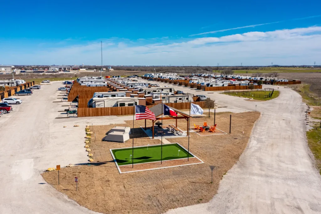 Aerial view of a fenced RV park with rows of parked trailers, a putting green, flagpoles, and covered picnic pavilion.